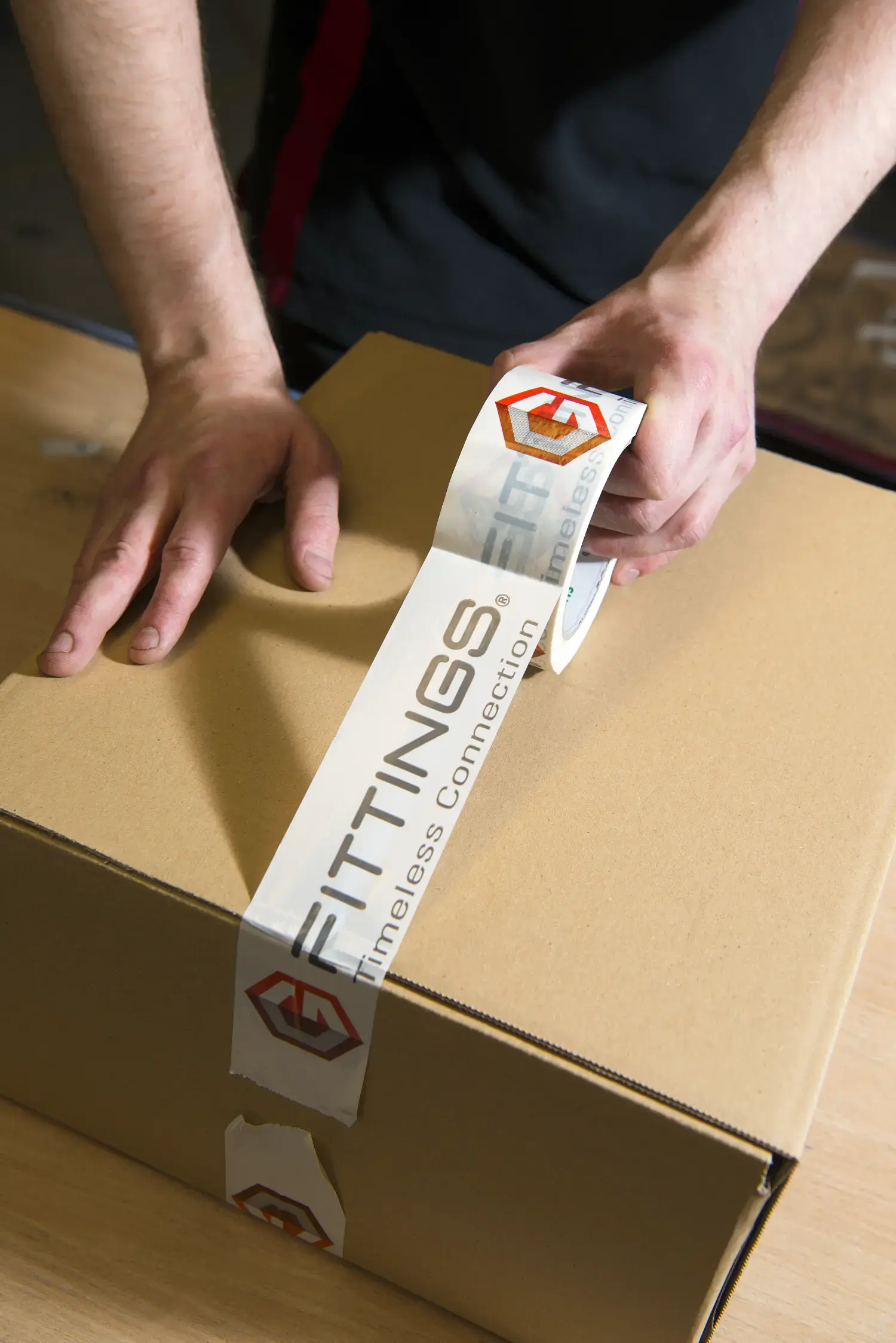 Eine Person verklebt einen Karton mit Hilfe einer Kleberolle, auf der ein G-Fittings Schriftzug zu lesen ist. Eine Person verklebt einen Karton mit Hilfe einer Kleberolle, auf der ein G-Fittings Schriftzug zu lesen ist.